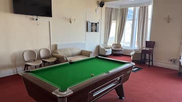 Een pooltafel in een kamer met een rood tapijt, beige banken en een flatscreen televisie aan de muur.