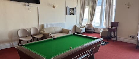 Ein Billardtisch in einem Raum mit rotem Teppich, beigen Sofas und einem an der Wand montierten Flachbildfernseher.