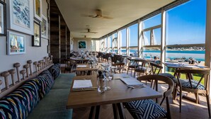 Un restaurante con una amplia zona de asientos tipo banco, mesas con sillas y grandes ventanales con vistas al mar.