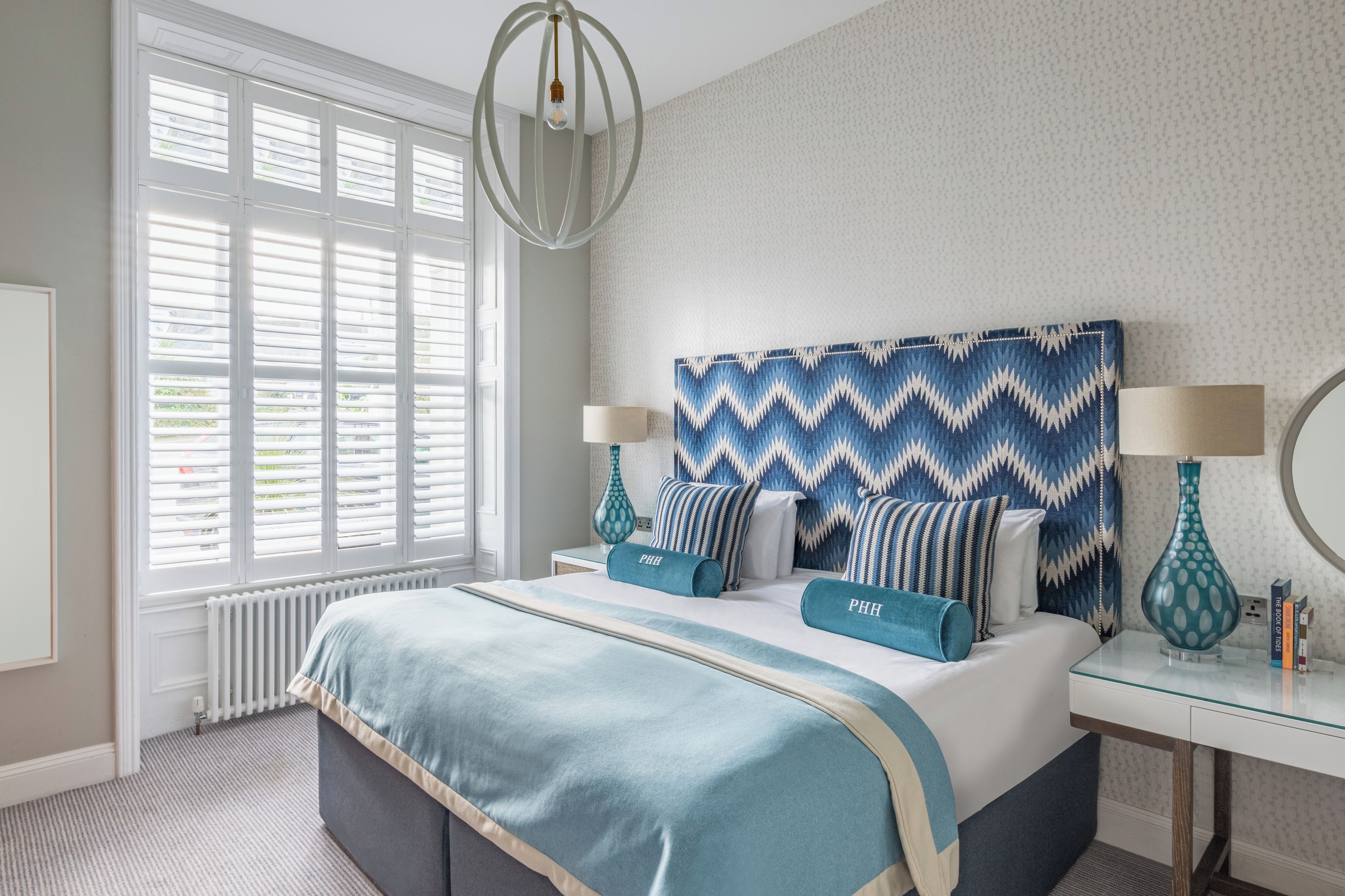 Une chambre à coucher avec un lit, des lampes de chevet, une fenêtre avec des stores et un mur décoré d’un motif.