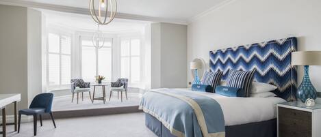 A bedroom with a large bed, a desk, and a chair. There is a window with blinds, a chandelier, and a patterned headboard.