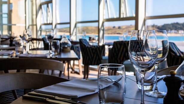 Un restaurante con mesas dispuestas para comer, cristalería transparente y vistas al mar a través de amplias ventanas.