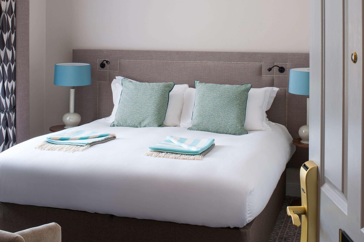 A hotel room with a large bed, two pillows, two bedside lamps, a nightstand, a chair, and a door with a number plate.