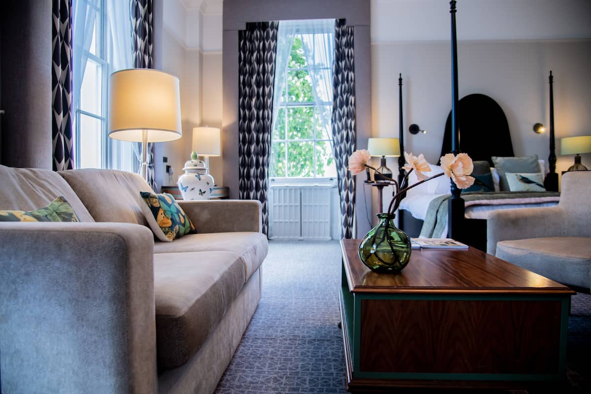 A hotel room with a sofa, a wooden coffee table with a vase, a bed with a headboard, and a window with curtains.