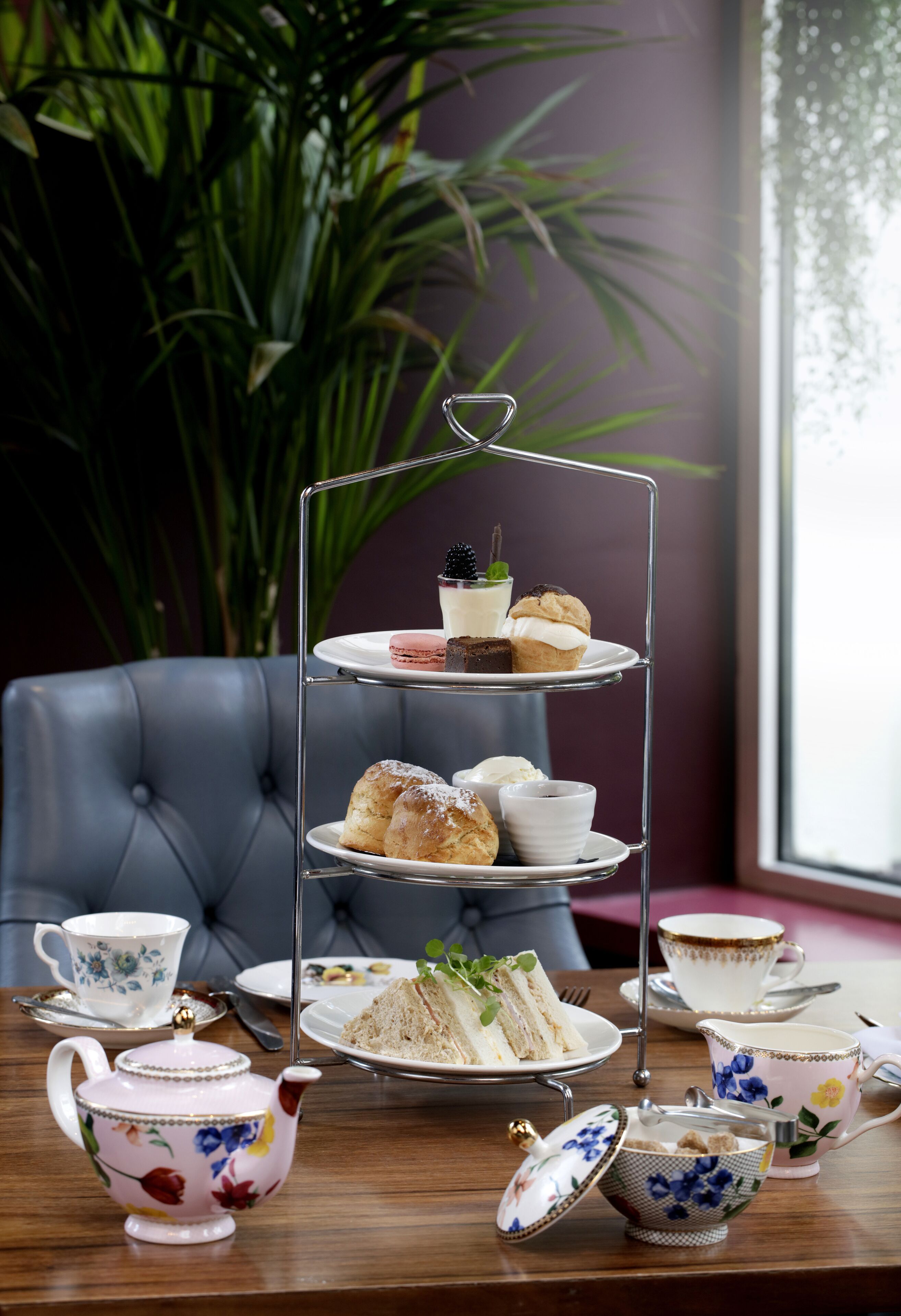 A three-tiered tea service stand with pastries and sandwiches, surrounded by floral teacups and a teapot on a wooden table.