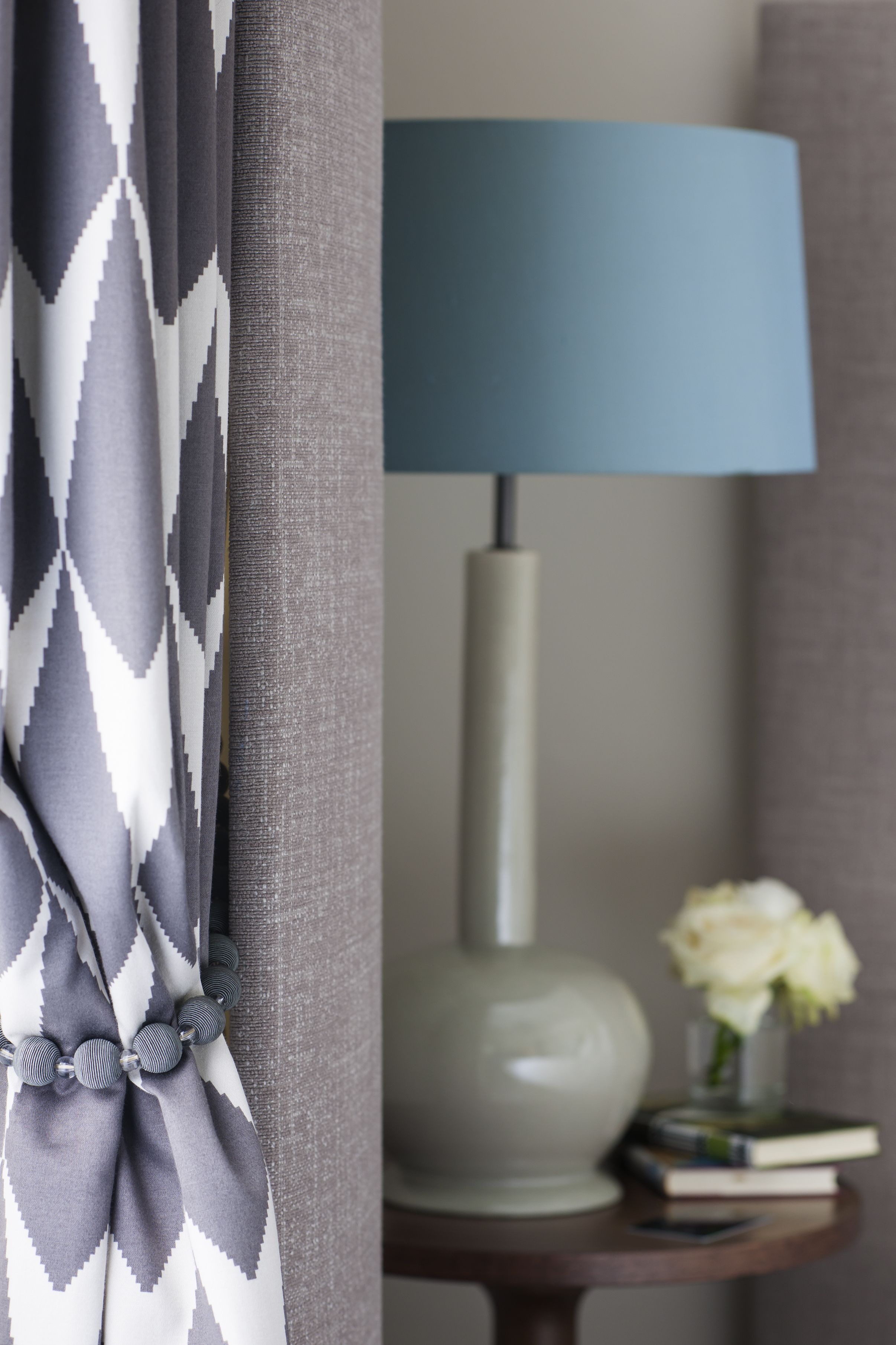 A room with a grey and white patterned curtain, a blue lamp on a side table, and a vase with white flowers.