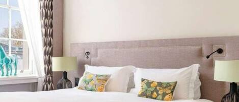 Una cama bien tendida con sábanas blancas y dos almohadones decorativos, una lámpara de noche y una ventana con cortinas.