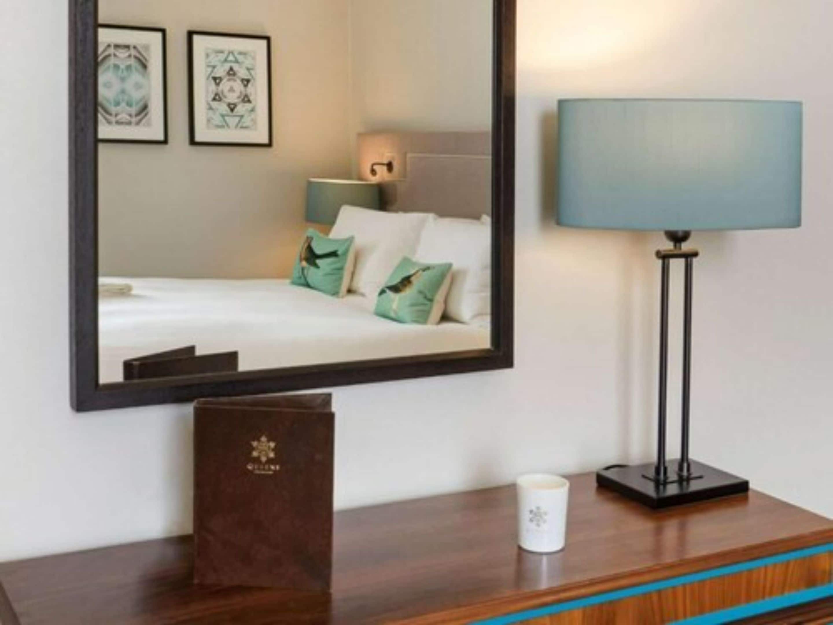 A hotel room with a bed, a bedside lamp, a framed mirror, and a small table with a mug.
