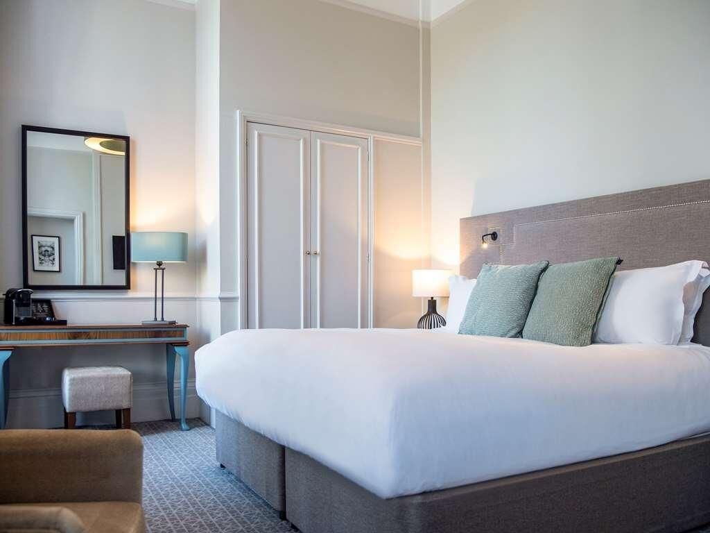 A hotel room with a bed, a desk, a chair, a lamp, and a mirror.