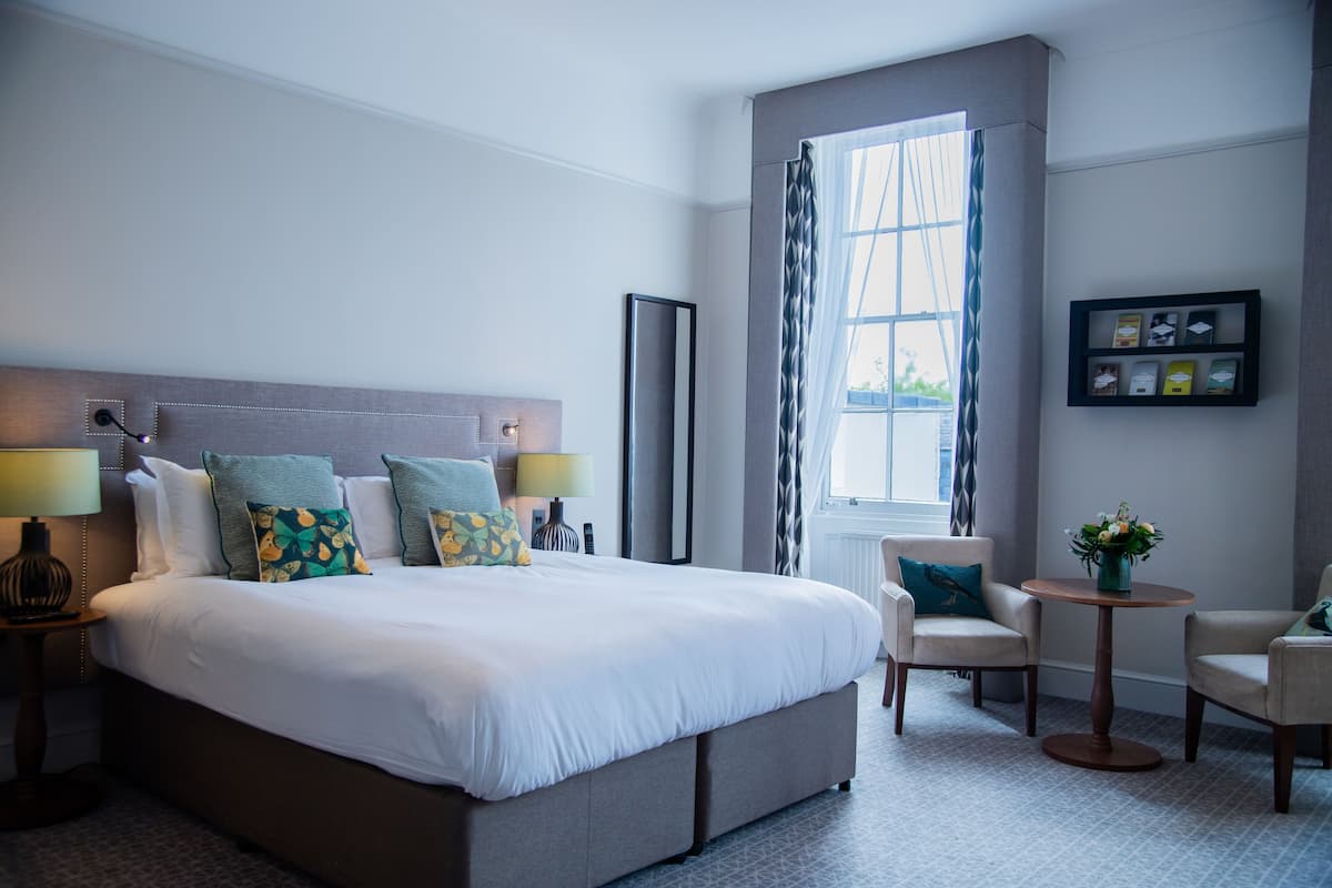 A hotel room with a large bed, two bedside tables, a small round table, two armchairs, and a window with curtains.