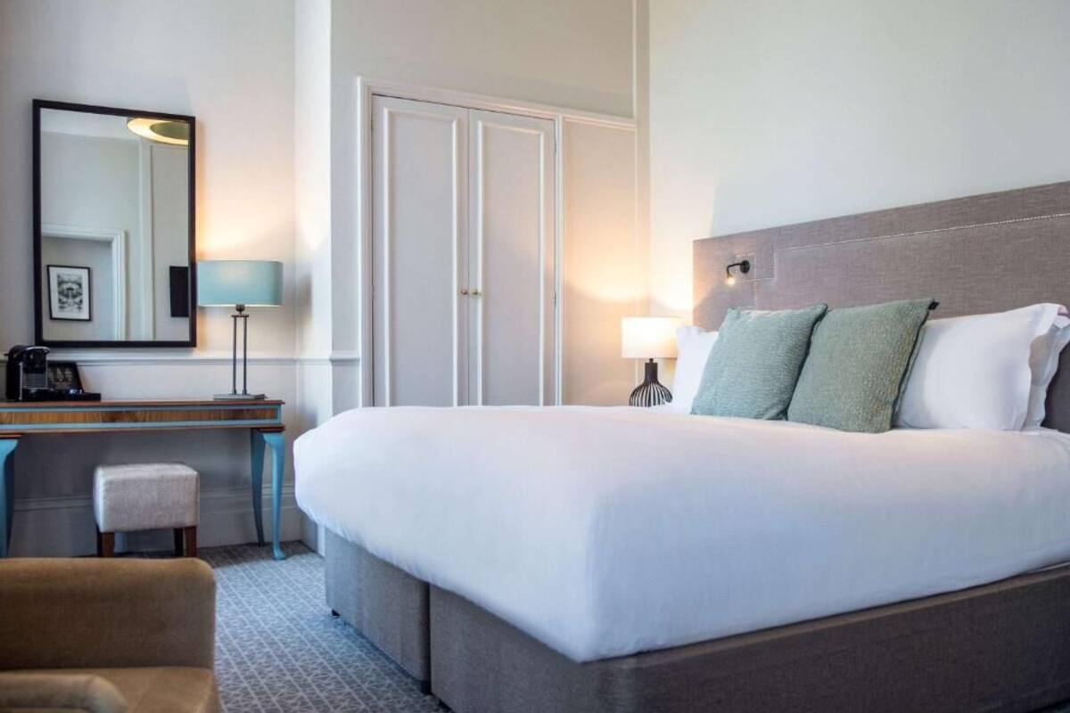 A hotel room with a large bed, a desk with a chair, a lamp, and a wardrobe.