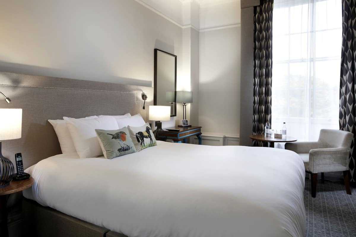 A hotel room with a large bed, bedside lamps, a small table, a chair, and a window with curtains.