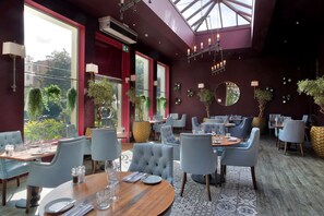 A restaurant with round tables, cushioned chairs, and a view of greenery through large windows. - Queens Hotel Cheltenham (Cheltenham)