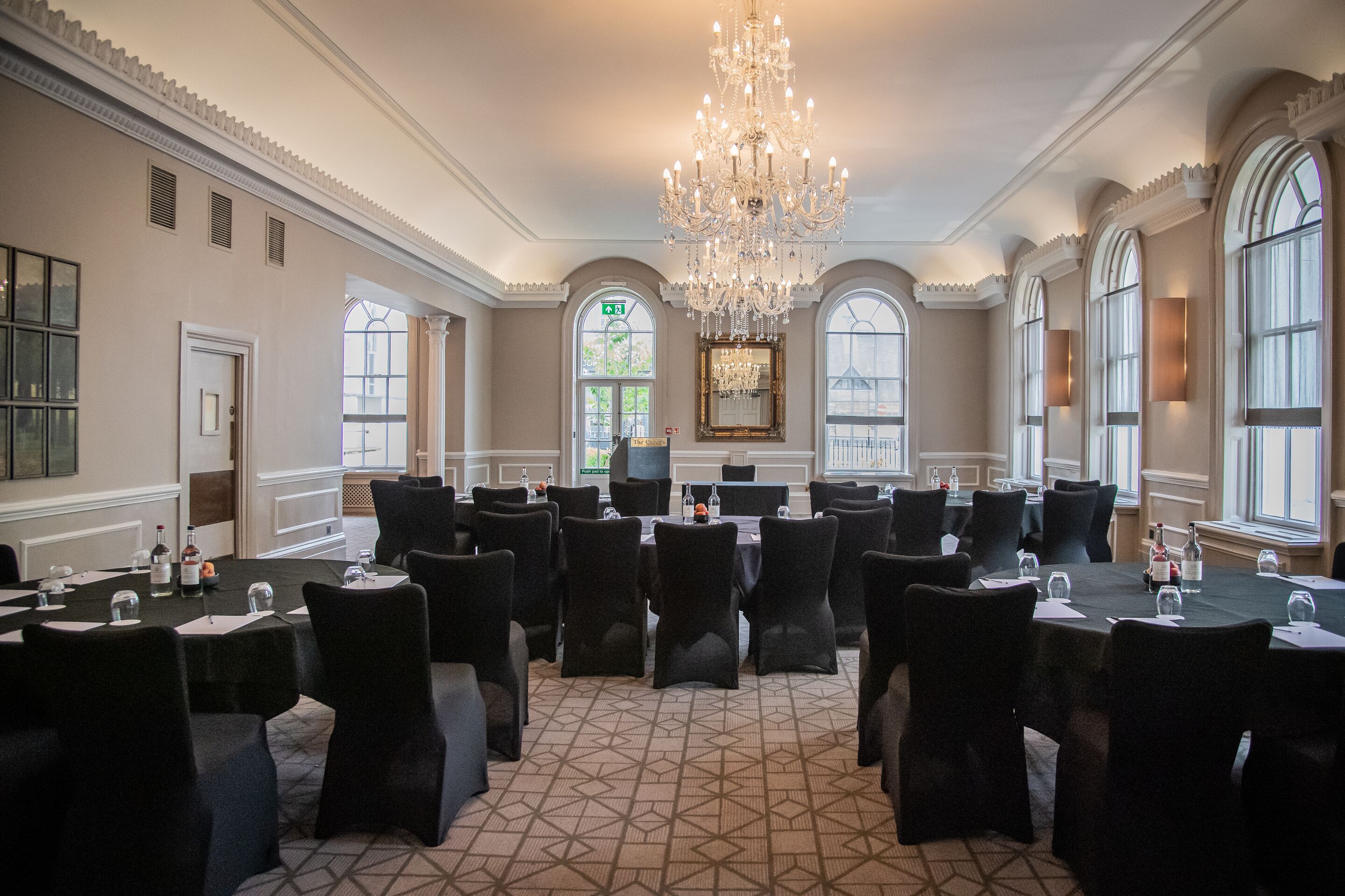 A formal meeting room with a large chandelier, multiple tables with chairs, and large windows with arched tops.