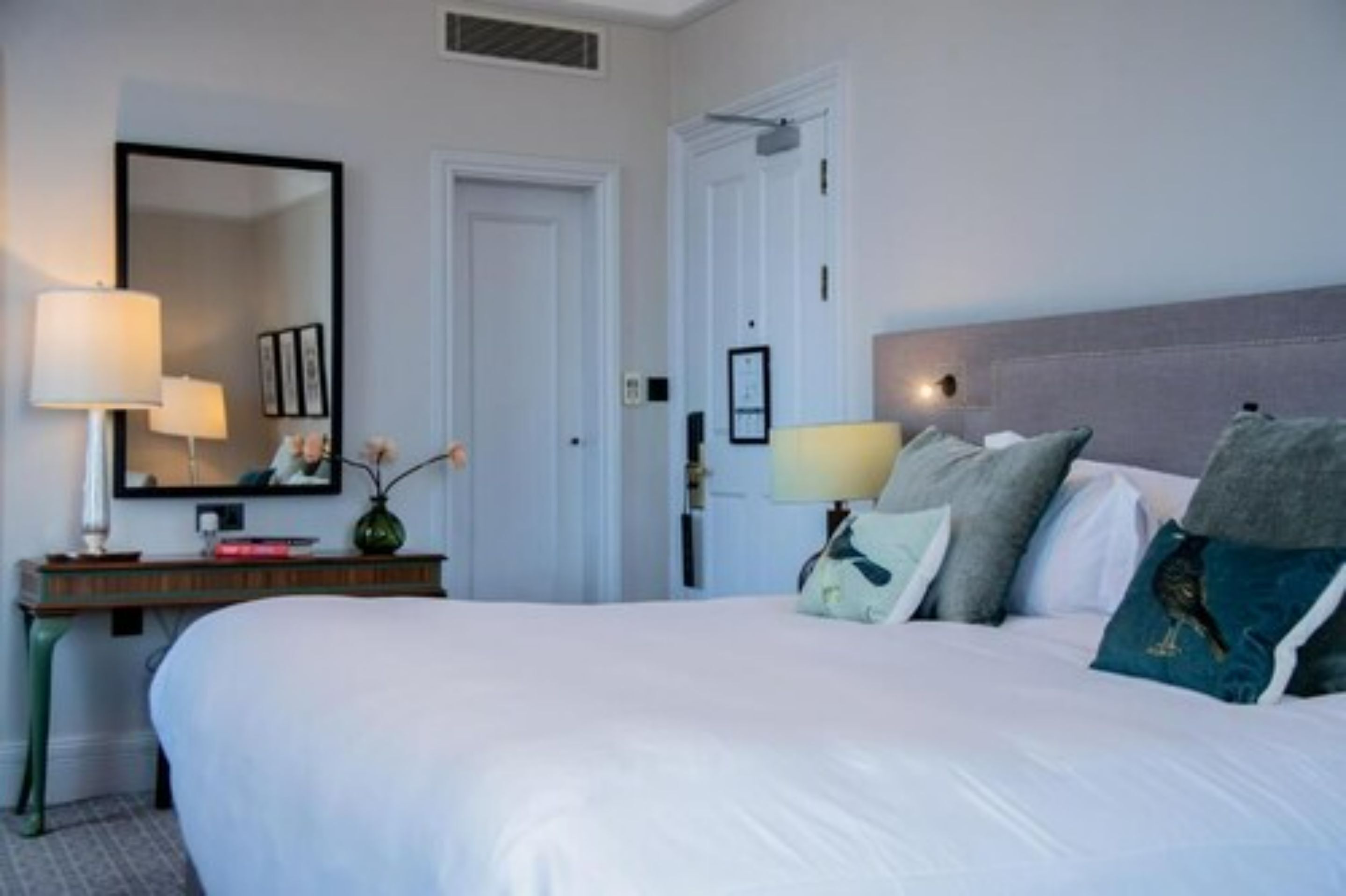 A hotel room with a large bed, a bedside table with a lamp, a mirror, and a door.