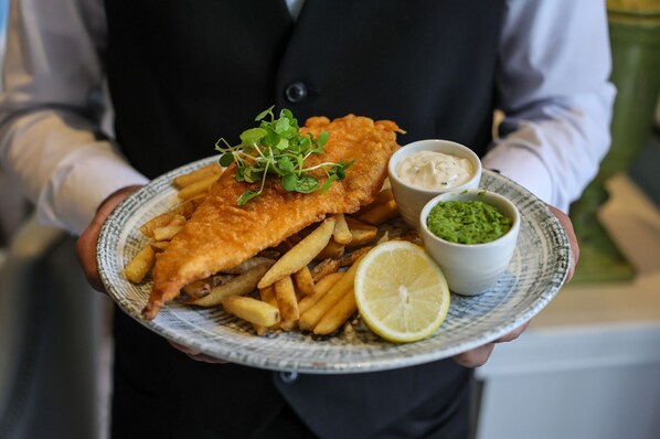 A person holding a plate with fish and chips, accompanied by two dipping sauces and a lemon wedge. - Queens Hotel Cheltenham (Cheltenham)