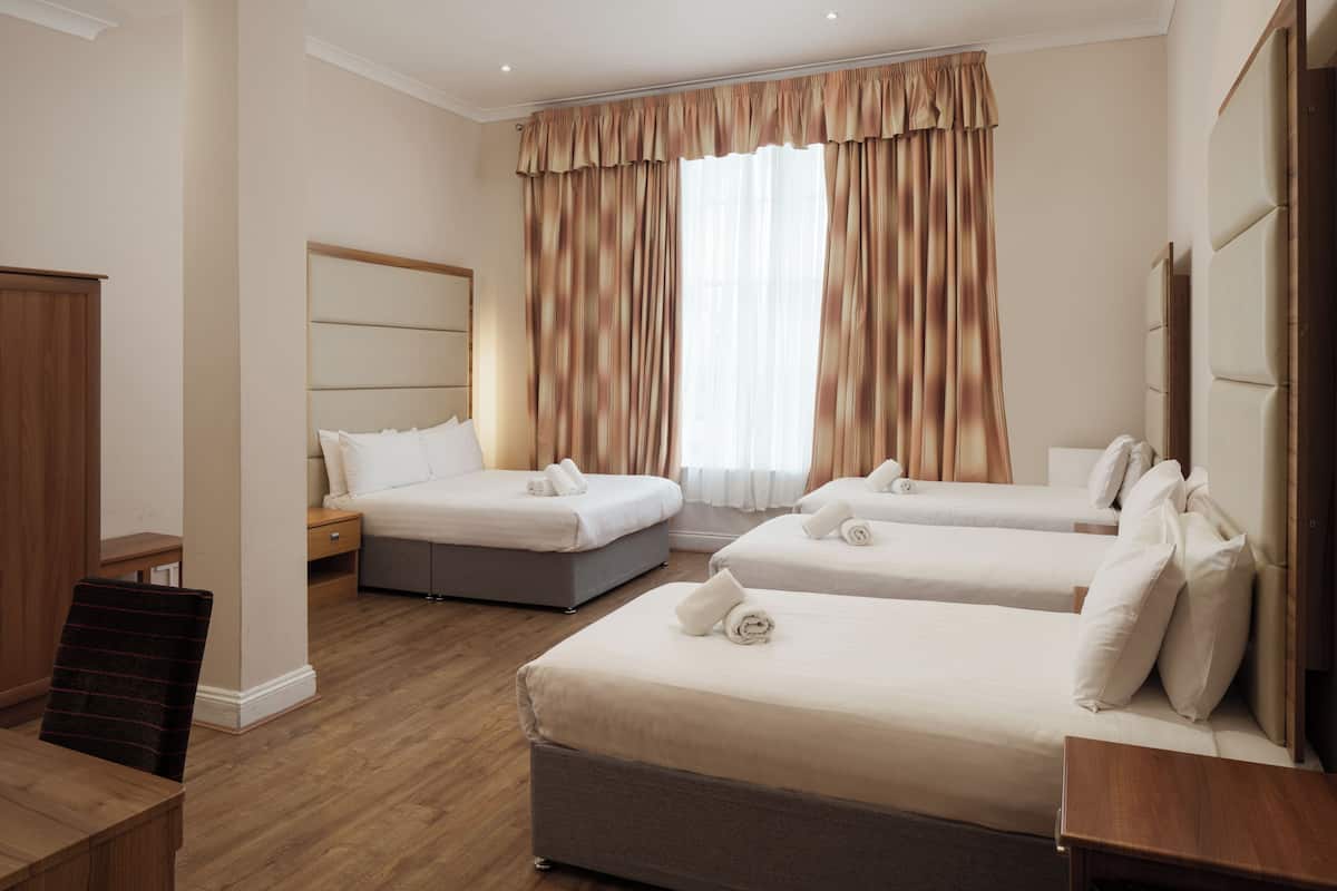 A hotel room with three beds, a wooden desk, and a large window with curtains.