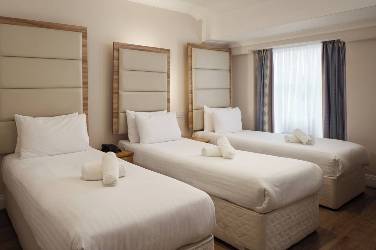 A hotel room with three beds, each with a headboard, a bedside table, and a rolled towel on each bed.