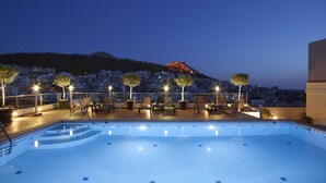 Uma área de piscina no terraço com espreguiçadeiras e árvores em vasos, com vista para a paisagem urbana ao entardecer.