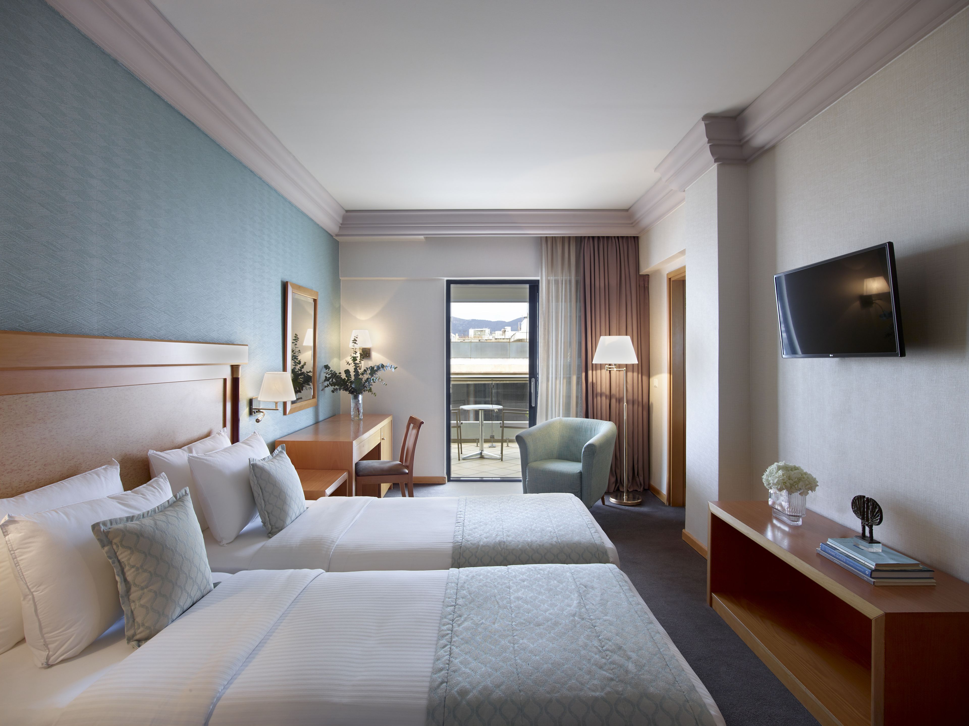 A hotel room with two beds, a desk, a chair, a TV, and a view of the city.
