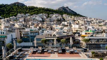 A rooftop pool area with lounge chairs and umbrellas, overlooking a densely packed urban area with buildings and a hill in the background.