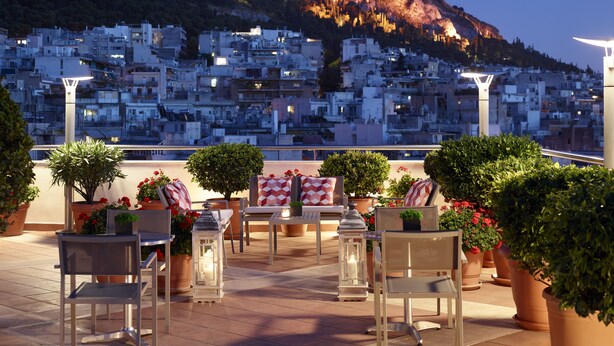 Um terraço ao ar livre com mesas e cadeiras, plantas em vasos e vista para uma cidade densamente construída numa encosta ao entardecer.