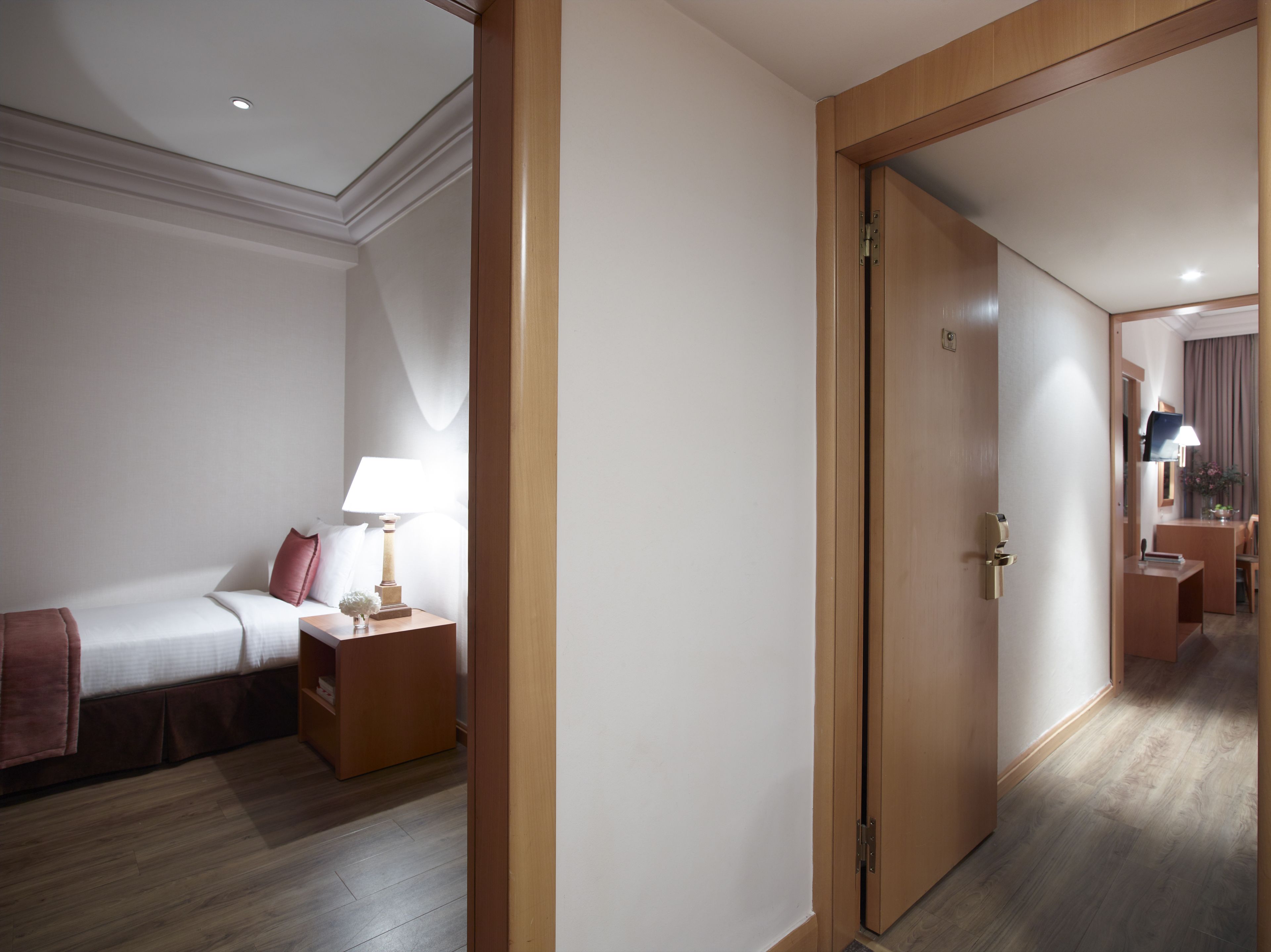 A hotel room with a bed, bedside table, lamp, and a door leading to another room.