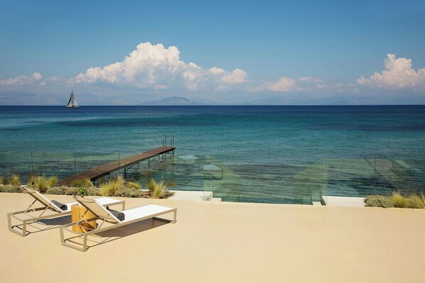 A beach with clear blue water, a sailboat, and a wooden deck with lounge chairs.