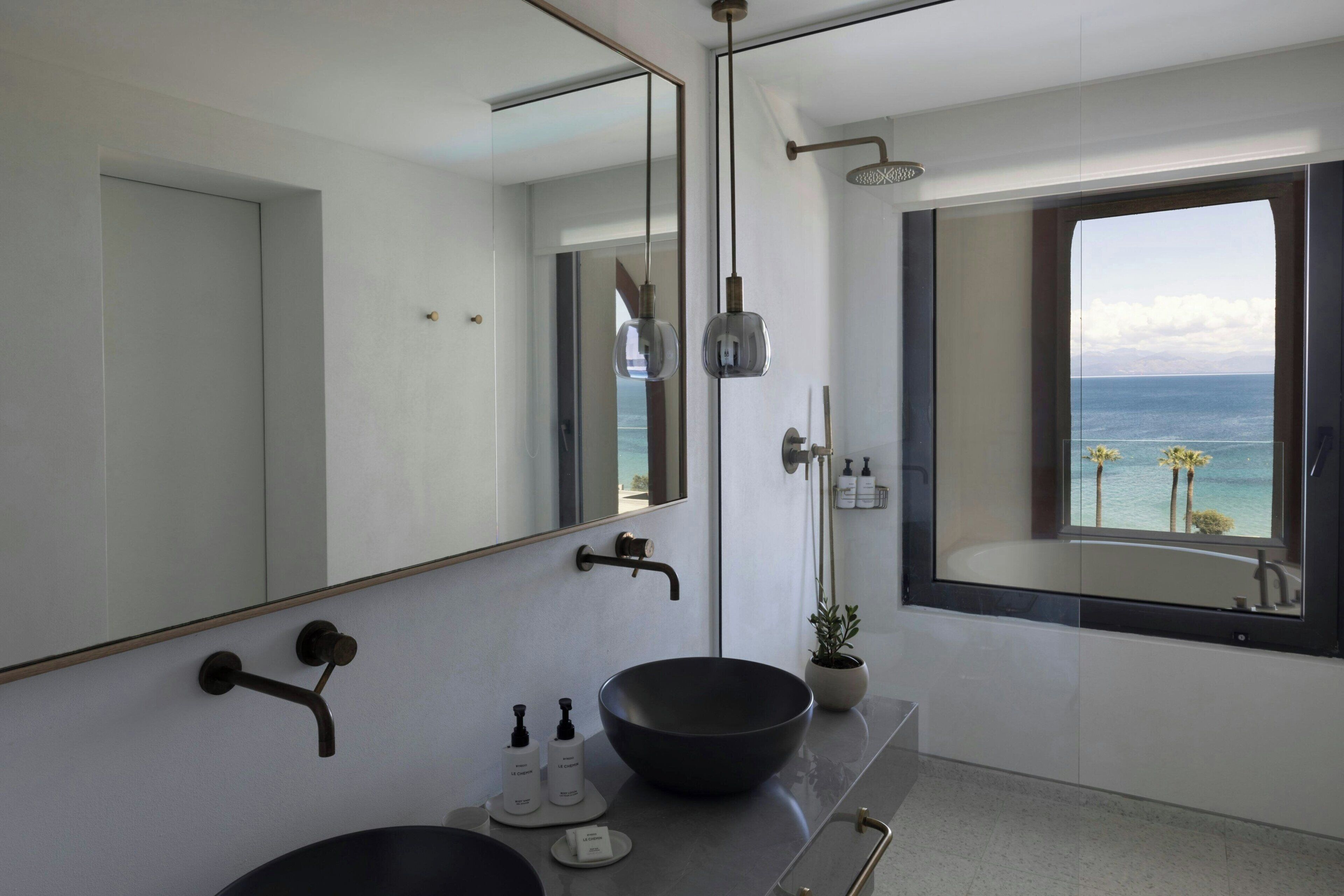 A modern bathroom with a large mirror, a black vessel sink, a shower with a handheld showerhead, and a view of the sea through a window.