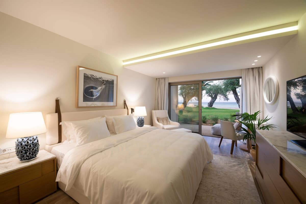 A modern hotel room with a large bed, two bedside tables with lamps, a desk with a chair, and a large window offering a view of the outdoors.