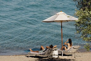 Two people relaxing on beach chairs under an umbrella by the water.