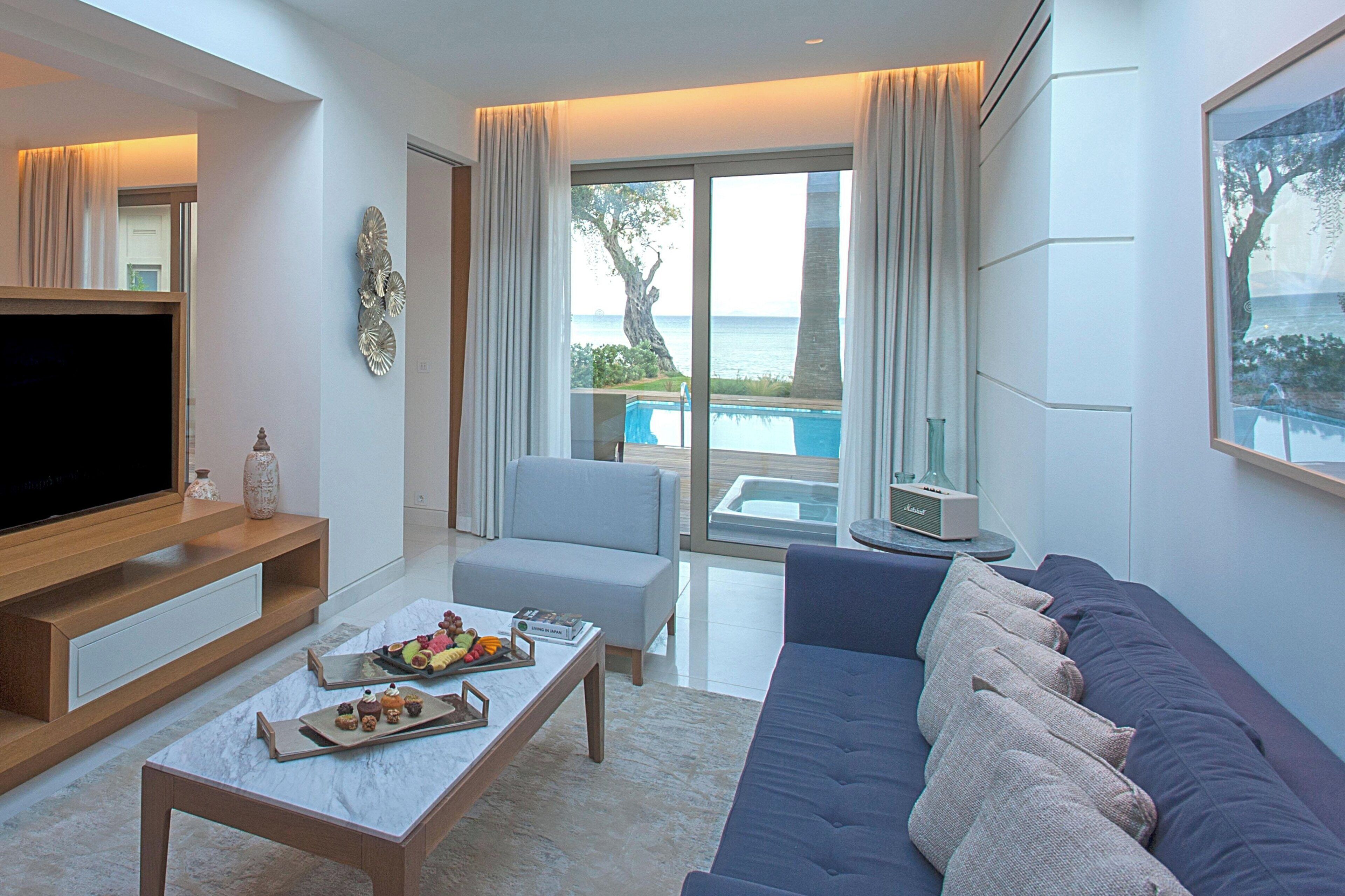 A modern living room with a flat-screen TV, a coffee table with snacks, a sofa, and a large window with a view of the sea.