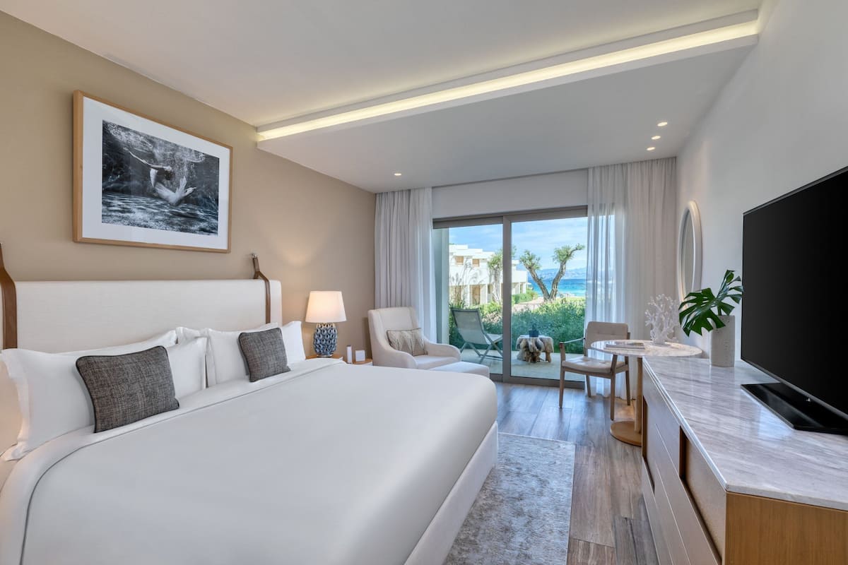 A modern hotel room with a large bed, a flat-screen TV, a dining area, and a balcony with a view of the sea.