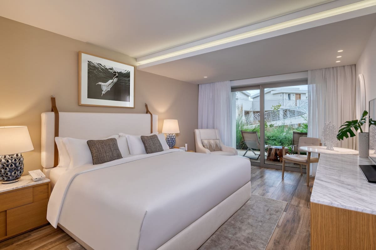 A hotel room with a large bed, bedside lamps, a desk with a chair, and a window with a view of greenery.