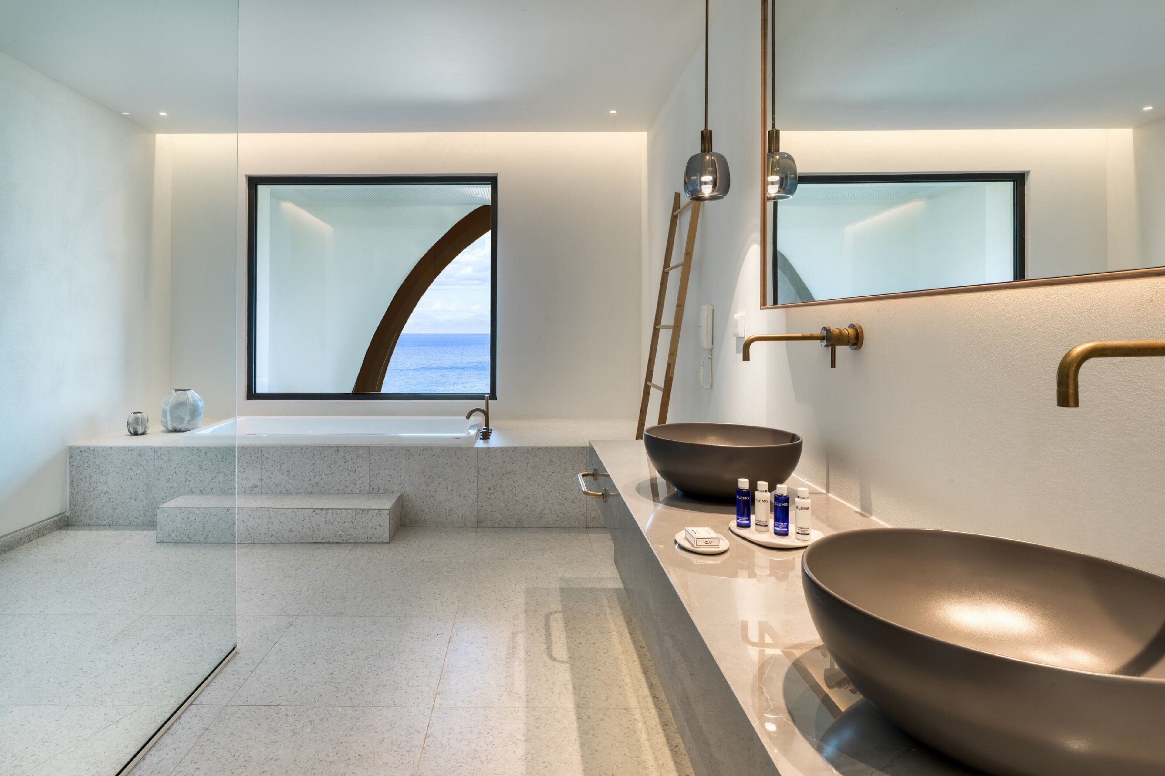 A modern bathroom with a large mirror, a bathtub, a large bowl sink, and a view of the sea.