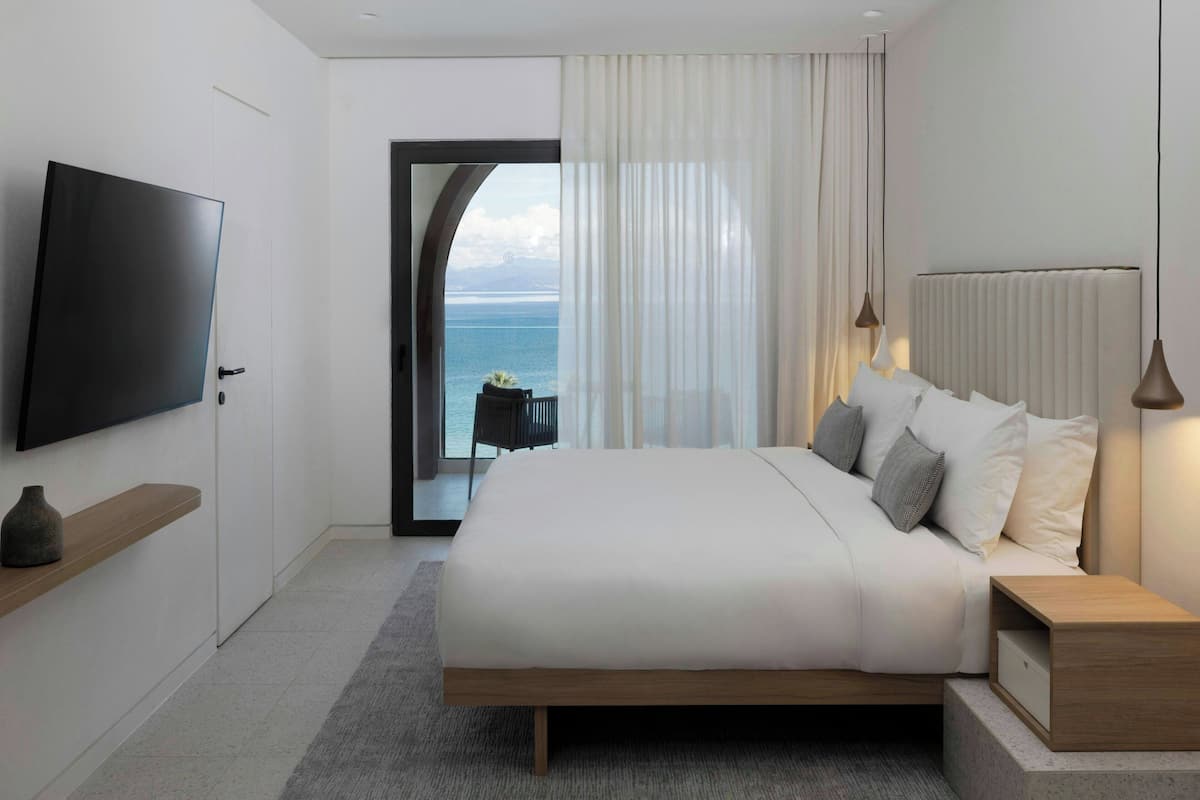 A modern bedroom with a bed, bedside tables, a TV, and a view of the sea.