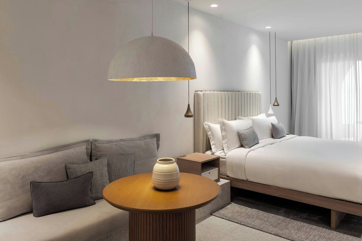 A modern hotel room with a sofa, a round wooden table, a bed, and a large pendant light.