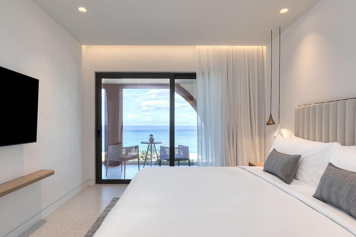 A modern hotel room with a large bed, a TV mounted on the wall, and a balcony with a view of the sea.