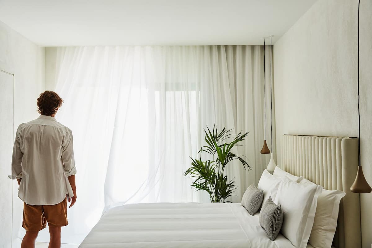 A bedroom with a bed, curtains, a potted plant, and a person standing by the window.