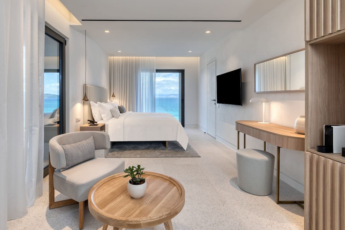 A modern hotel room with a large bed, a seating area with a table, a TV, and a view of the sea.