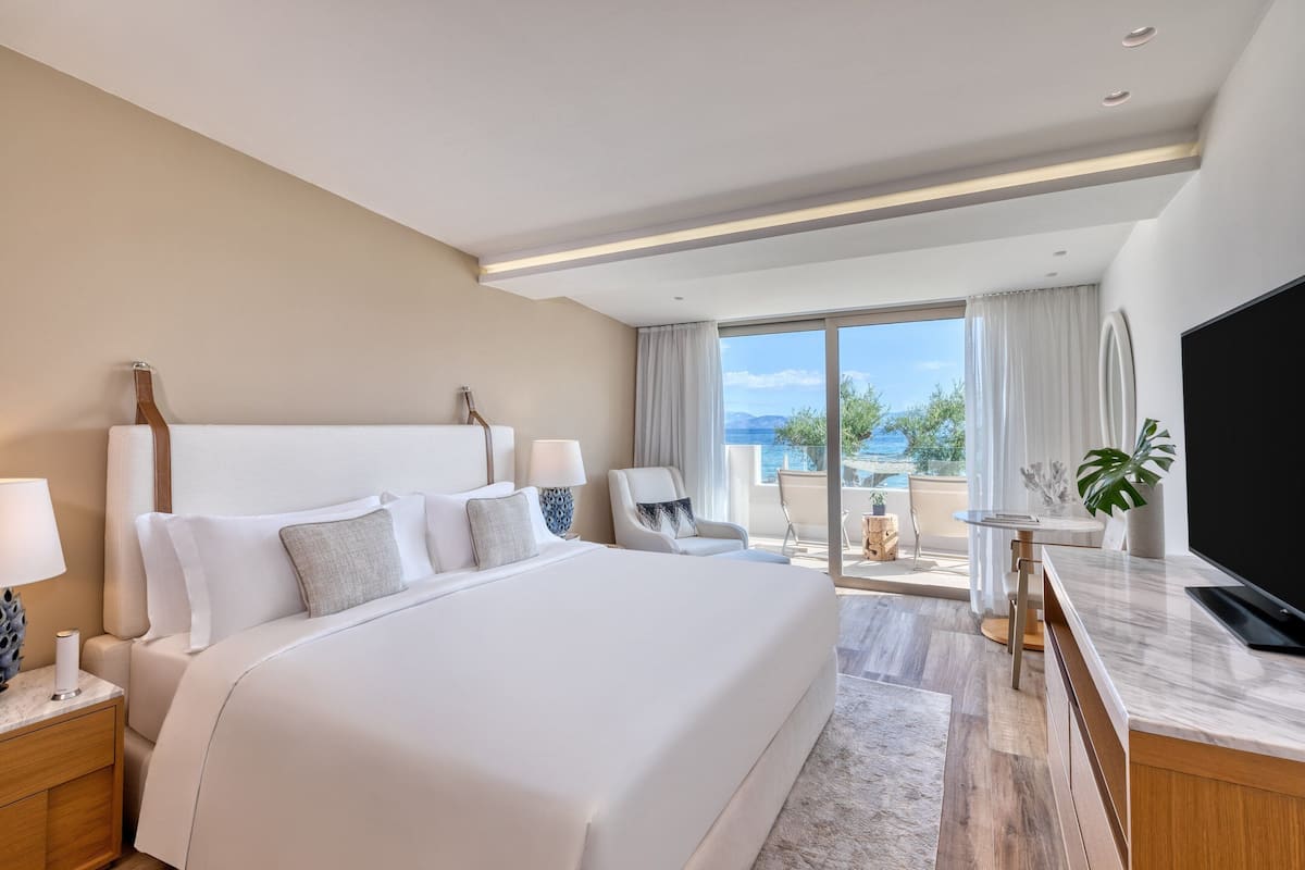 A modern hotel room with a large bed, a desk with a flat-screen TV, a chair, and a view of the outdoors.