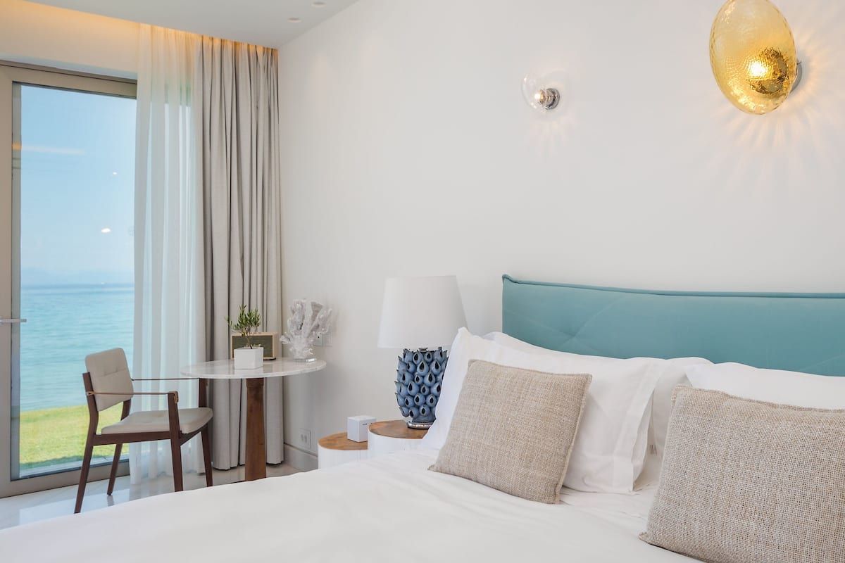 A hotel room with a large bed, a view of the sea, a round table with a chair, and a nightstand with a lamp and decorative items.