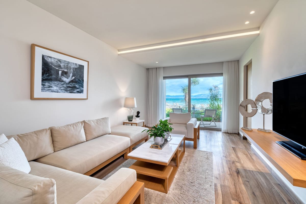 A modern living room with a sofa, coffee table, and TV. There is a large window with a view of the sea.