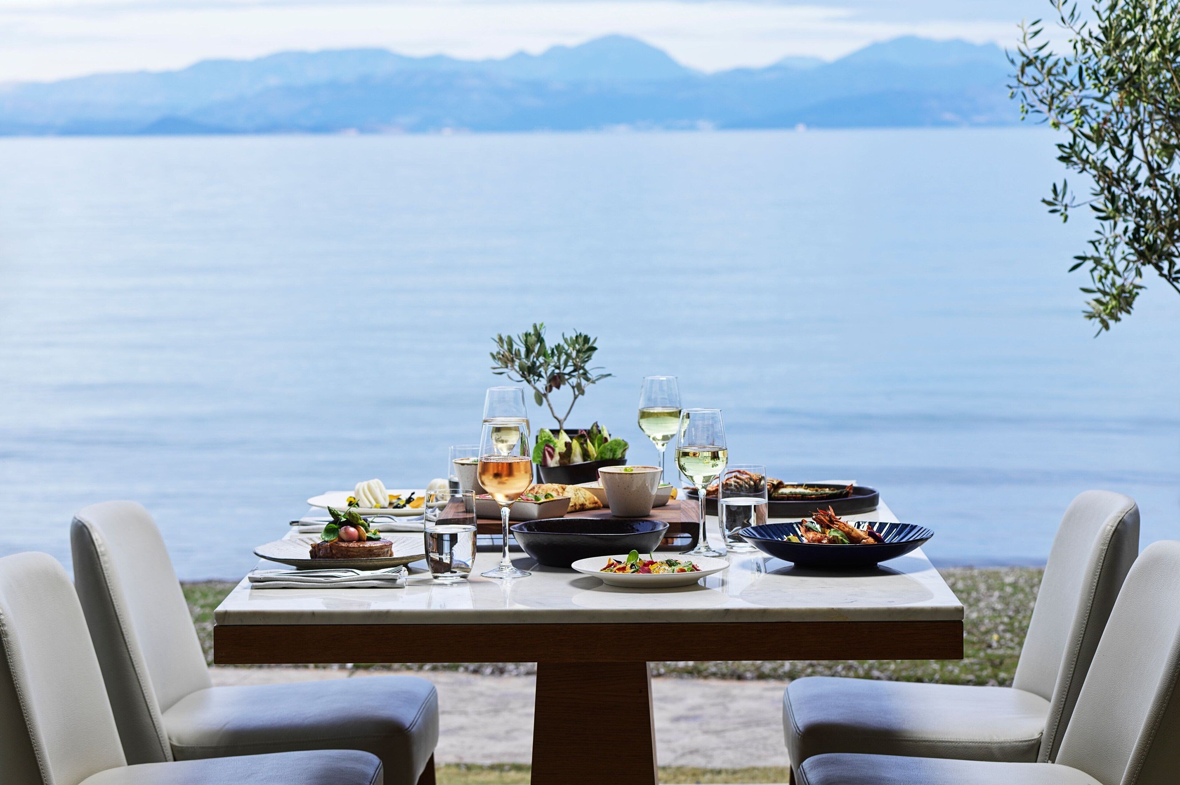 A dining table set with various dishes and glasses, overlooking a body of water with mountains in the background.