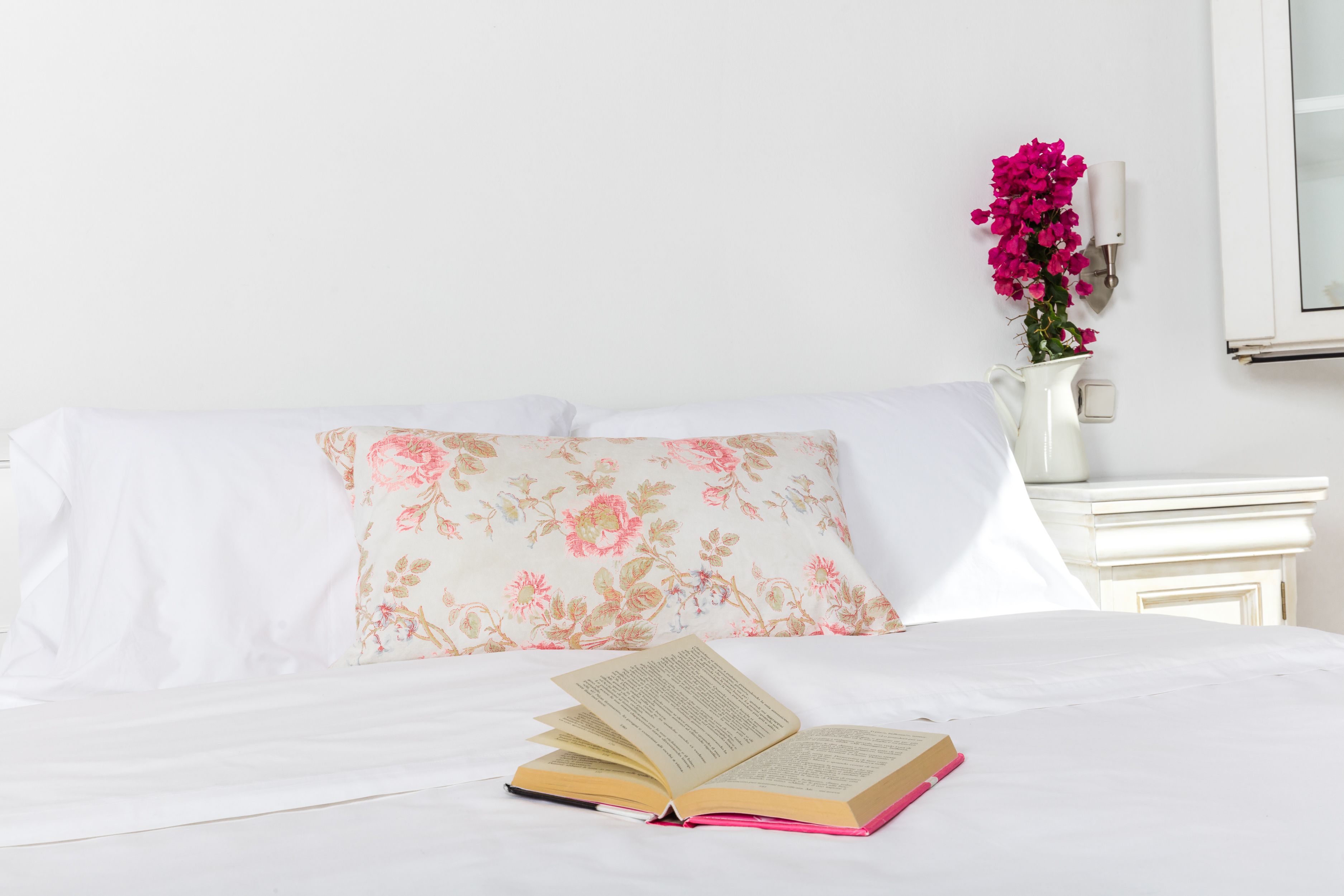 A neatly made bed with a floral pillow, a white bedside table with a vase of pink flowers, and an open book on the bed.