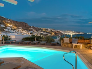 Zona de piscina con sillas reclinables y vistas a un pueblo costero al anochecer.