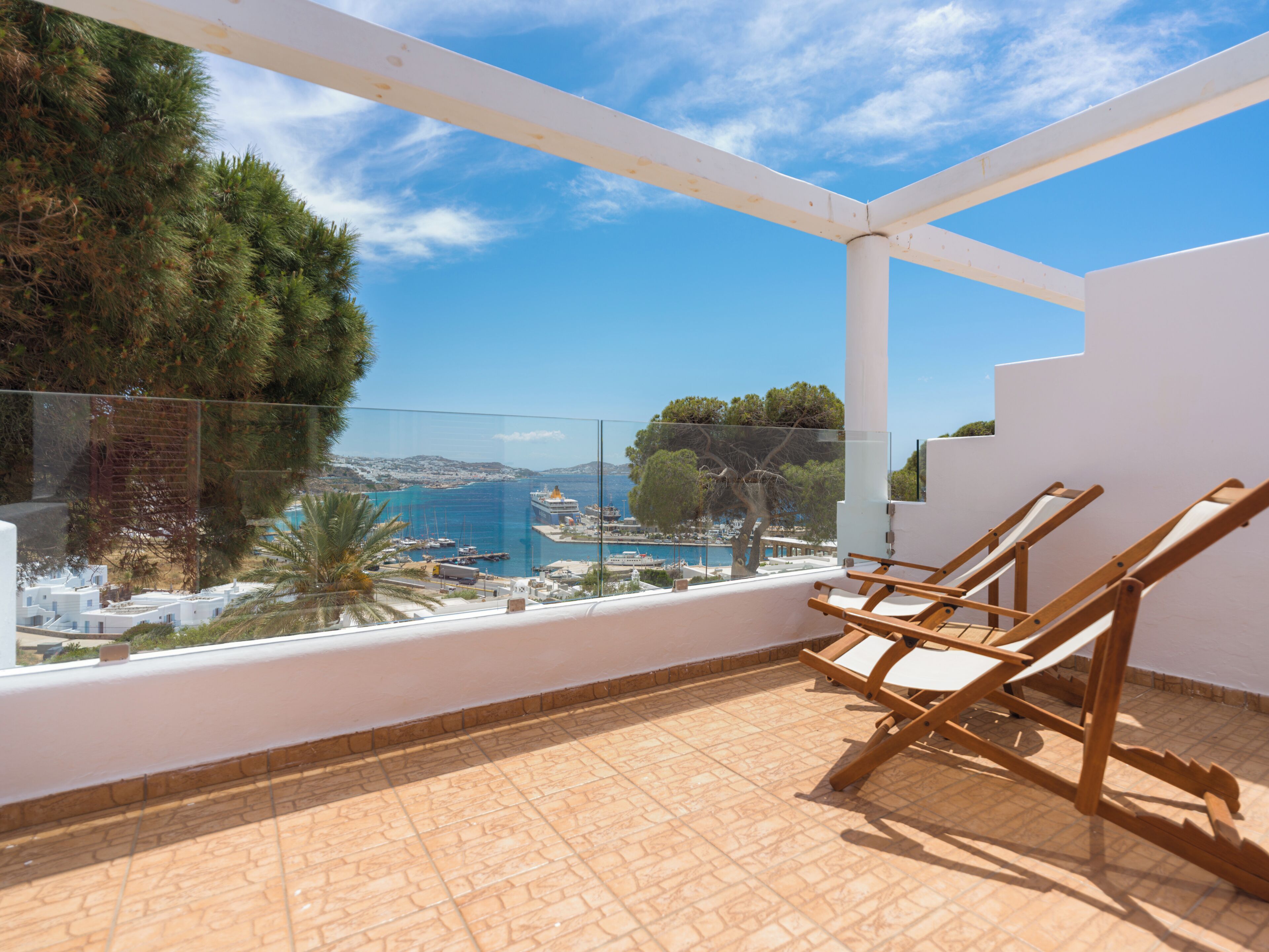A balcony with a view of the sea, wooden chairs, and a tiled floor.
