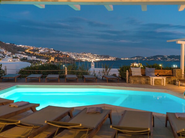 A pool area with lounge chairs and a view of a coastal town at dusk.