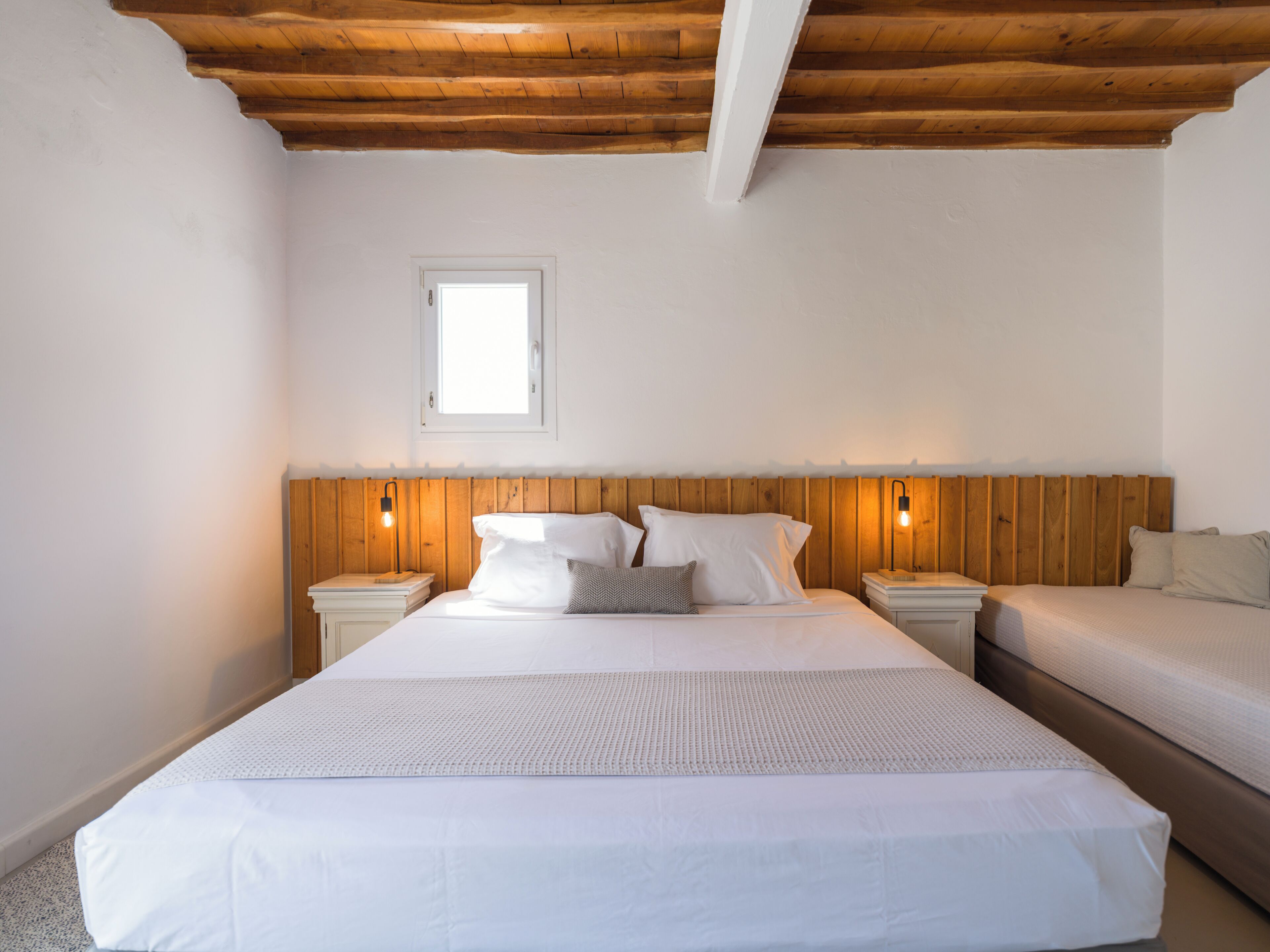 A bedroom with a large bed, two bedside tables, a wooden headboard, a window, and a wall-mounted light fixture.
