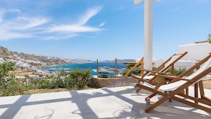 Una terraza frente al mar con sillones de madera, una pérgola blanca y vistas al océano y edificios lejanos.
